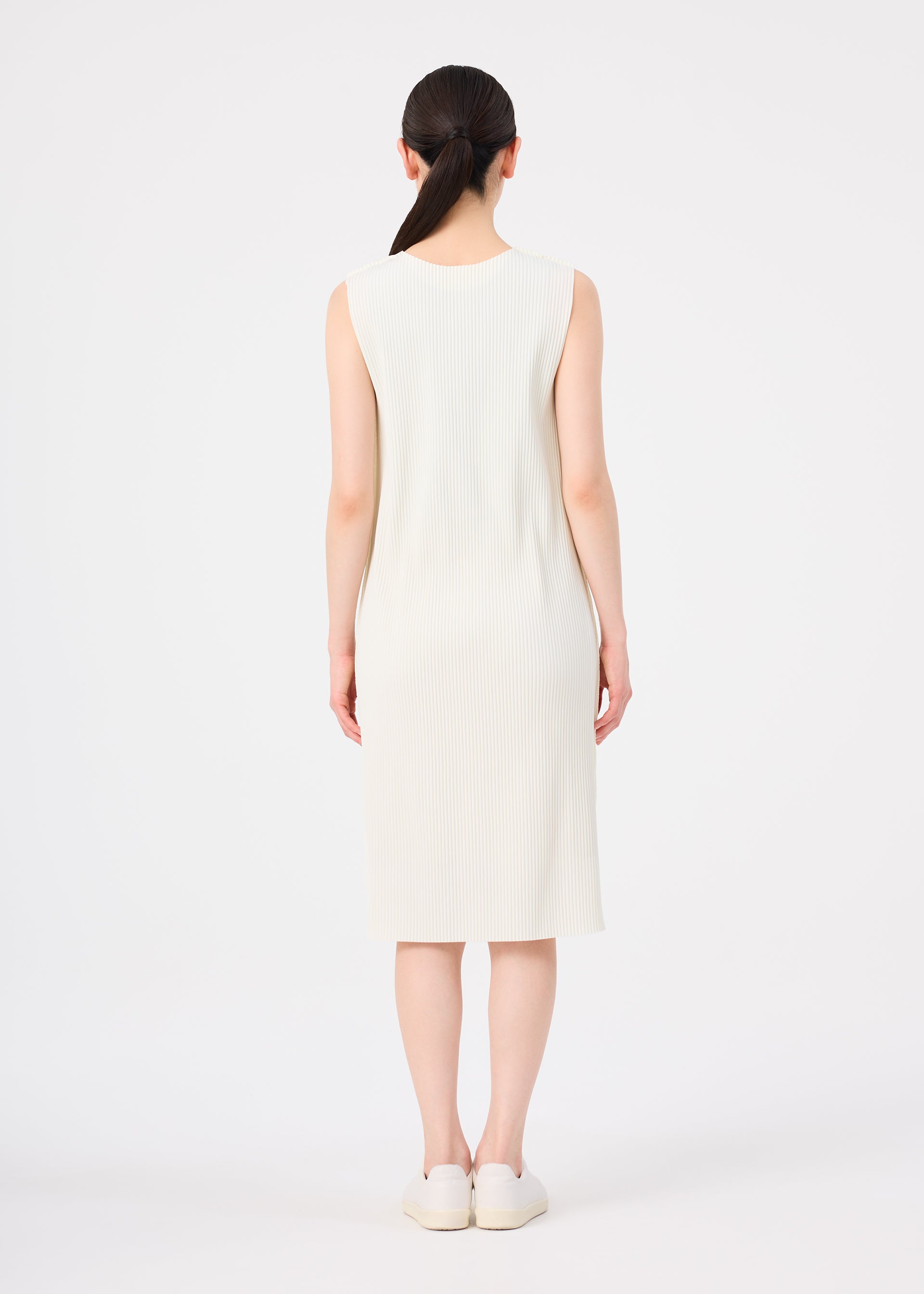 Zoomed view A woman with dark hair in a low ponytail stands facing away, wearing a sleeveless, knee-length cream FINE KNIT PLEATS dress from me ISSEY MIYAKE and white slip-on shoes against a plain white background.