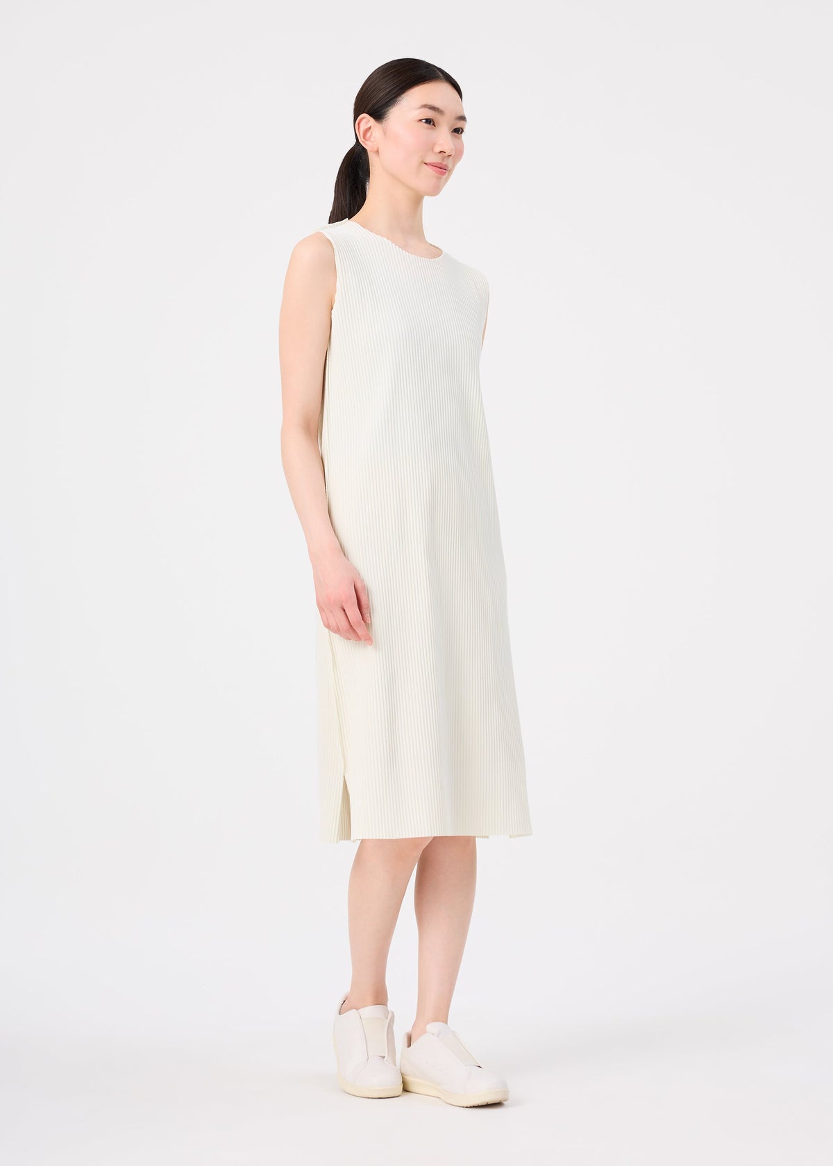 A woman with straight, dark hair pulled back wears the me ISSEY MIYAKE FINE KNIT PLEATS sleeveless, off-white knee-length dress with subtle fine knit pleats and white slip-on shoes, standing against a plain white background. view 7 of 7