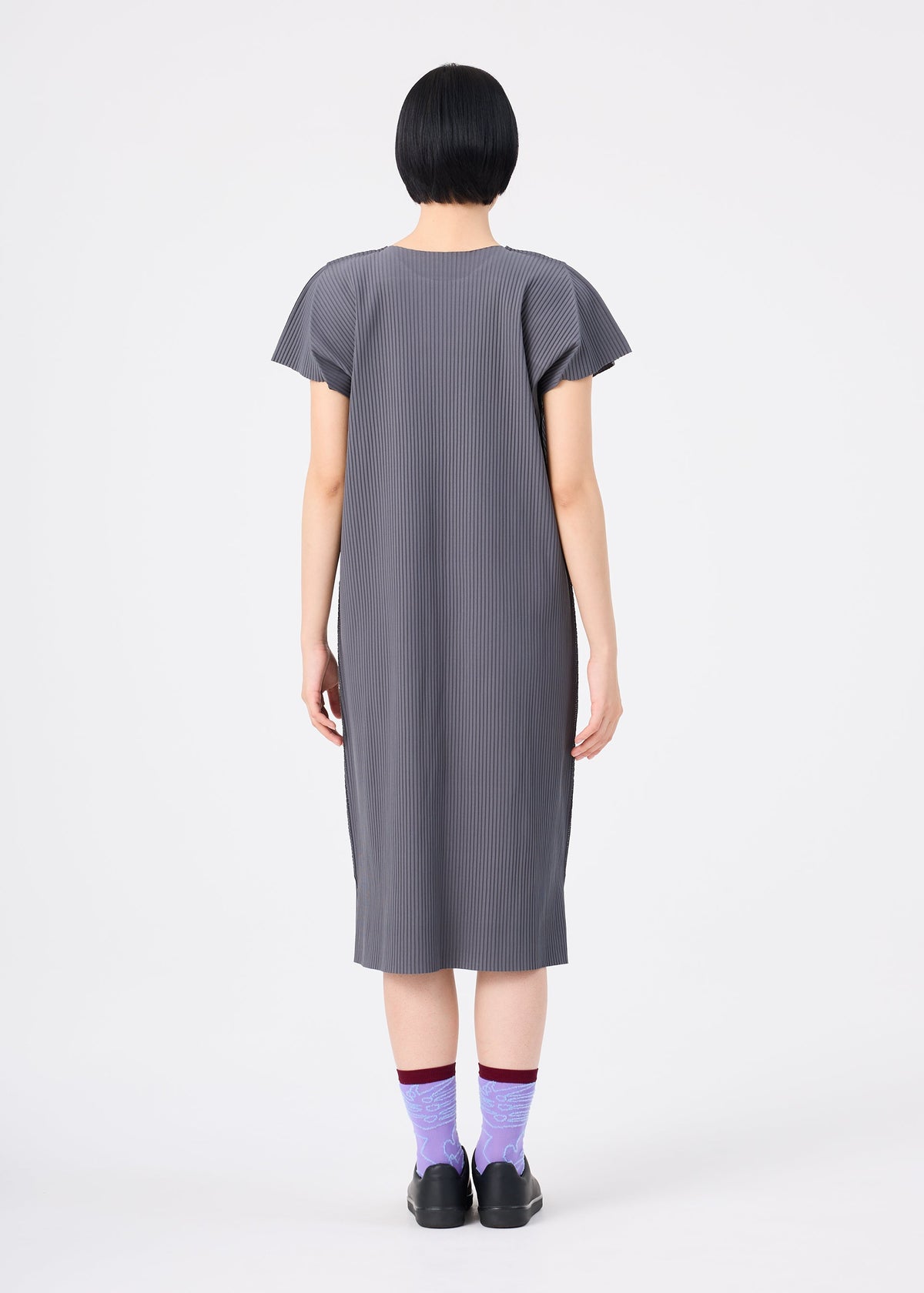 A person with short black hair, wearing the me ISSEY MIYAKE FINE KNIT PLEATS COLOR 2 gray, short-sleeved pleated dress and purple socks with black shoes, stands facing away against a plain white background. view 8 of 9