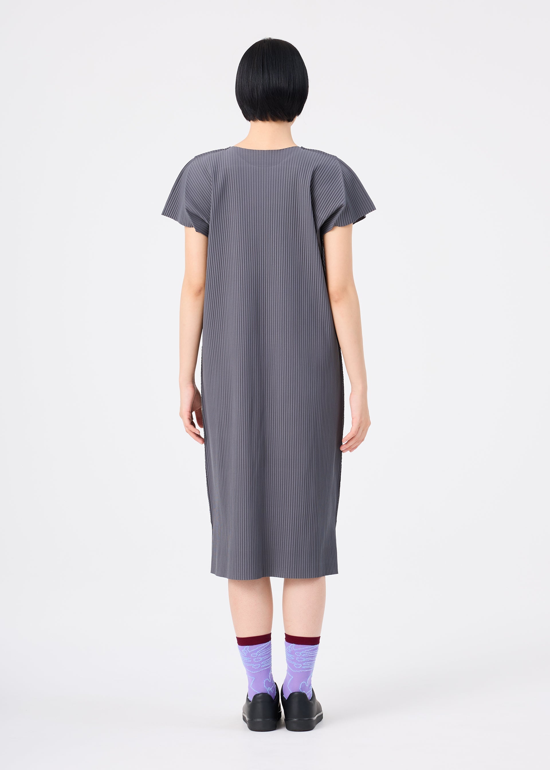 Zoomed view A person with short black hair, wearing the me ISSEY MIYAKE FINE KNIT PLEATS COLOR 2 gray, short-sleeved pleated dress and purple socks with black shoes, stands facing away against a plain white background.