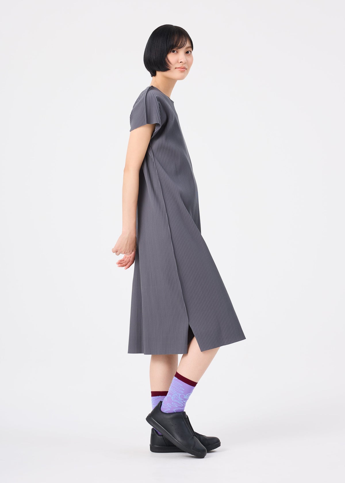 A woman with short black hair stands sideways against a plain white background, wearing the me ISSEY MIYAKE FINE KNIT PLEATS COLOR 2 dress in loose gray with short sleeves, paired with black ankle boots and purple patterned socks. view 9 of 9