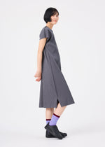 A woman with short black hair stands sideways against a plain white background, wearing the me ISSEY MIYAKE FINE KNIT PLEATS COLOR 2 dress in loose gray with short sleeves, paired with black ankle boots and purple patterned socks.