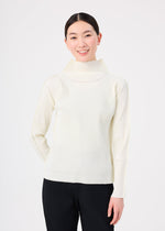 A woman with dark hair tied back smiles against a white background, wearing the FINE KNIT PLEATS top by me ISSEY MIYAKE—a light-colored, high-neck, long-sleeved piece with fine pleats—paired with dark pants.