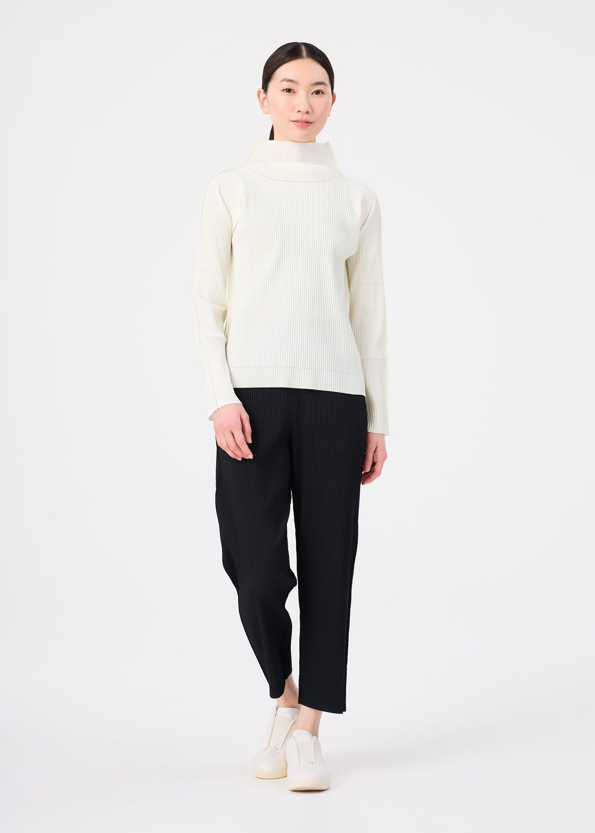 A woman with long dark hair in a low ponytail wears the FINE KNIT PLEATS by me ISSEY MIYAKE—cream high-neck top and loose black pants—paired with white slip-on shoes, standing against a plain white background. view 6 of 6