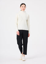 A woman with long dark hair in a low ponytail wears the FINE KNIT PLEATS by me ISSEY MIYAKE—cream high-neck top and loose black pants—paired with white slip-on shoes, standing against a plain white background.