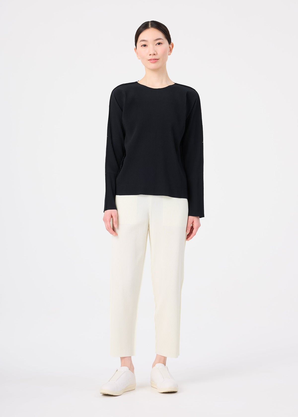 A person stands against a plain white background wearing the FINE KNIT PLEATS long-sleeved black top by me ISSEY MIYAKE, off-white straight-leg pants, and white shoes. Their straight dark hair is pulled back as they look forward neutrally. view 6 of 6