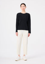 A person stands against a plain white background wearing the FINE KNIT PLEATS long-sleeved black top by me ISSEY MIYAKE, off-white straight-leg pants, and white shoes. Their straight dark hair is pulled back as they look forward neutrally.