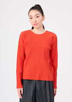 A woman with long dark hair in a ponytail wears the FINE KNIT PLEATS COLOR 2 top by me ISSEY MIYAKE—an orange, ribbed, long-sleeved round-neck top—paired with dark pleated pants. She stands against a plain white background, looking calmly at the camera.