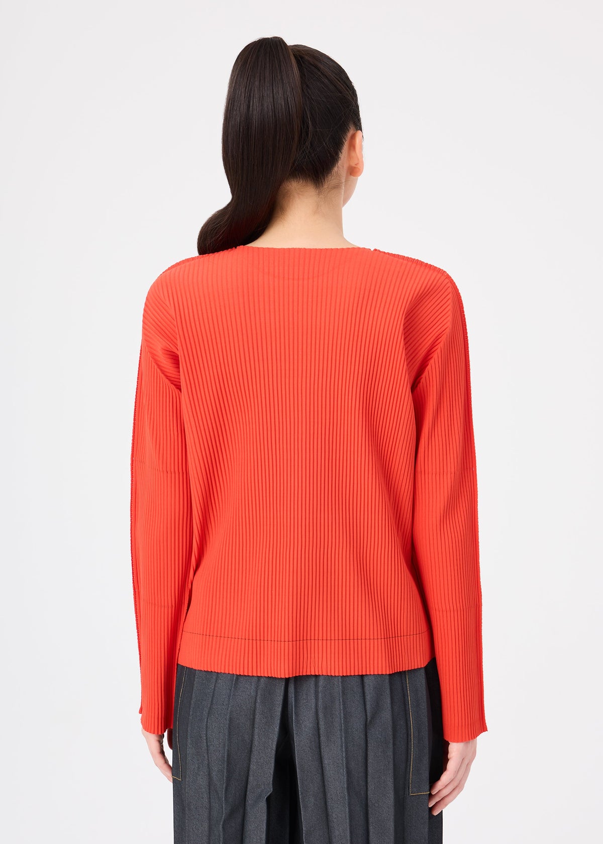 A person with long dark hair in a ponytail is seen from behind against a plain white background, wearing the me ISSEY MIYAKE FINE KNIT PLEATS COLOR 2 top in red and high-waisted gray pants. view 7 of 8