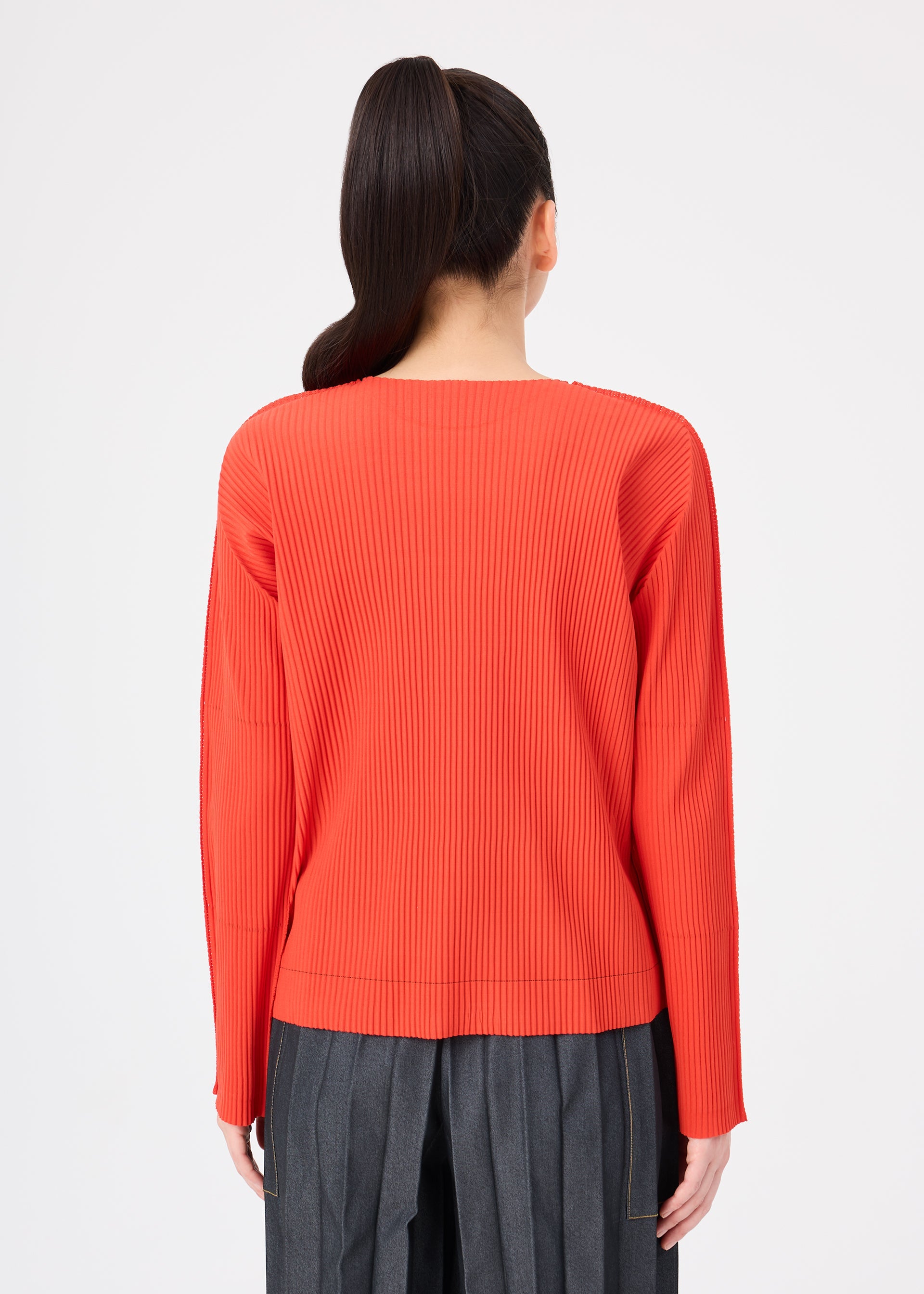 Zoomed view A person with long dark hair in a ponytail is seen from behind against a plain white background, wearing the me ISSEY MIYAKE FINE KNIT PLEATS COLOR 2 top in red and high-waisted gray pants.