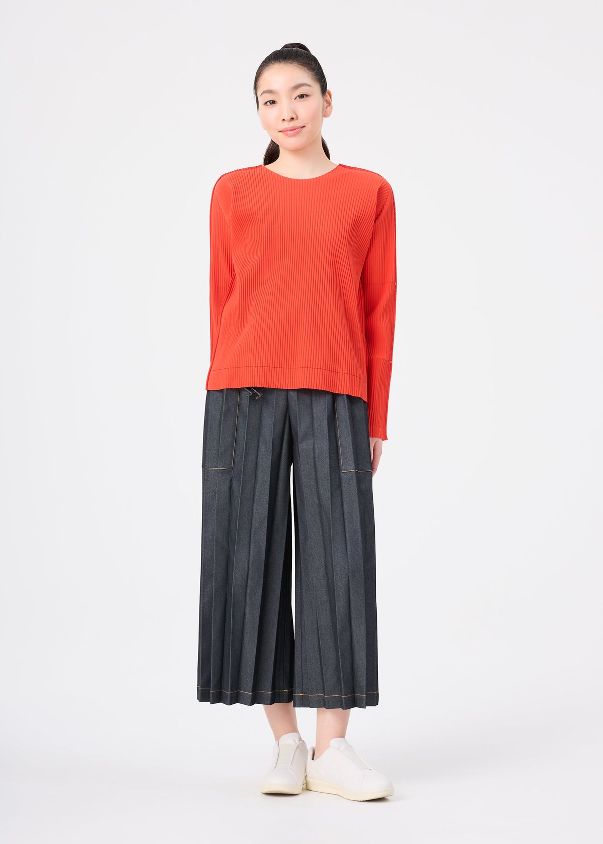 A woman stands against a white background, wearing a long-sleeved red round-neck top, the FINE KNIT PLEATS COLOR 2 wide-leg gray pleated pants by me ISSEY MIYAKE, and white sneakers. Her straight dark hair is tied back, and she smiles gently. view 8 of 8