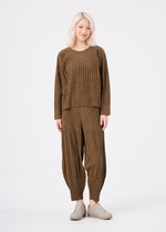 A person with light blonde hair wears the me ISSEY MIYAKE MIX FINE KNIT PLEATS set—loose long-sleeved top and matching wide-legged pants—with light slip-on shoes, standing against a plain white background.