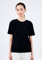 A woman with long dark hair tied back wears the me ISSEY MIYAKE FINE KNIT PLEATS black, round-neck short-sleeved top and white pants, standing against a plain white background and facing forward with a neutral expression.