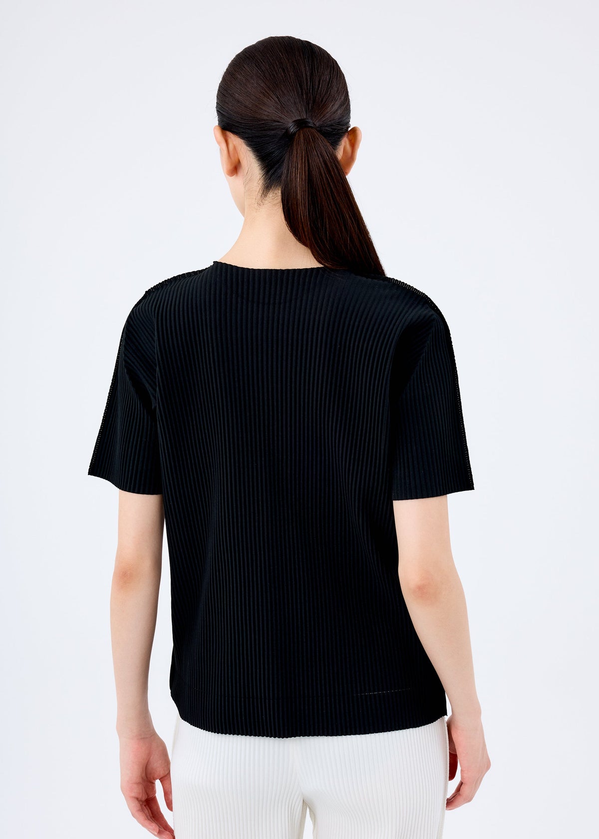 A person with long dark hair in a low ponytail is seen from behind wearing the me ISSEY MIYAKE FINE KNIT PLEATS black short-sleeved top and white pants against a plain light background. view 5 of 6