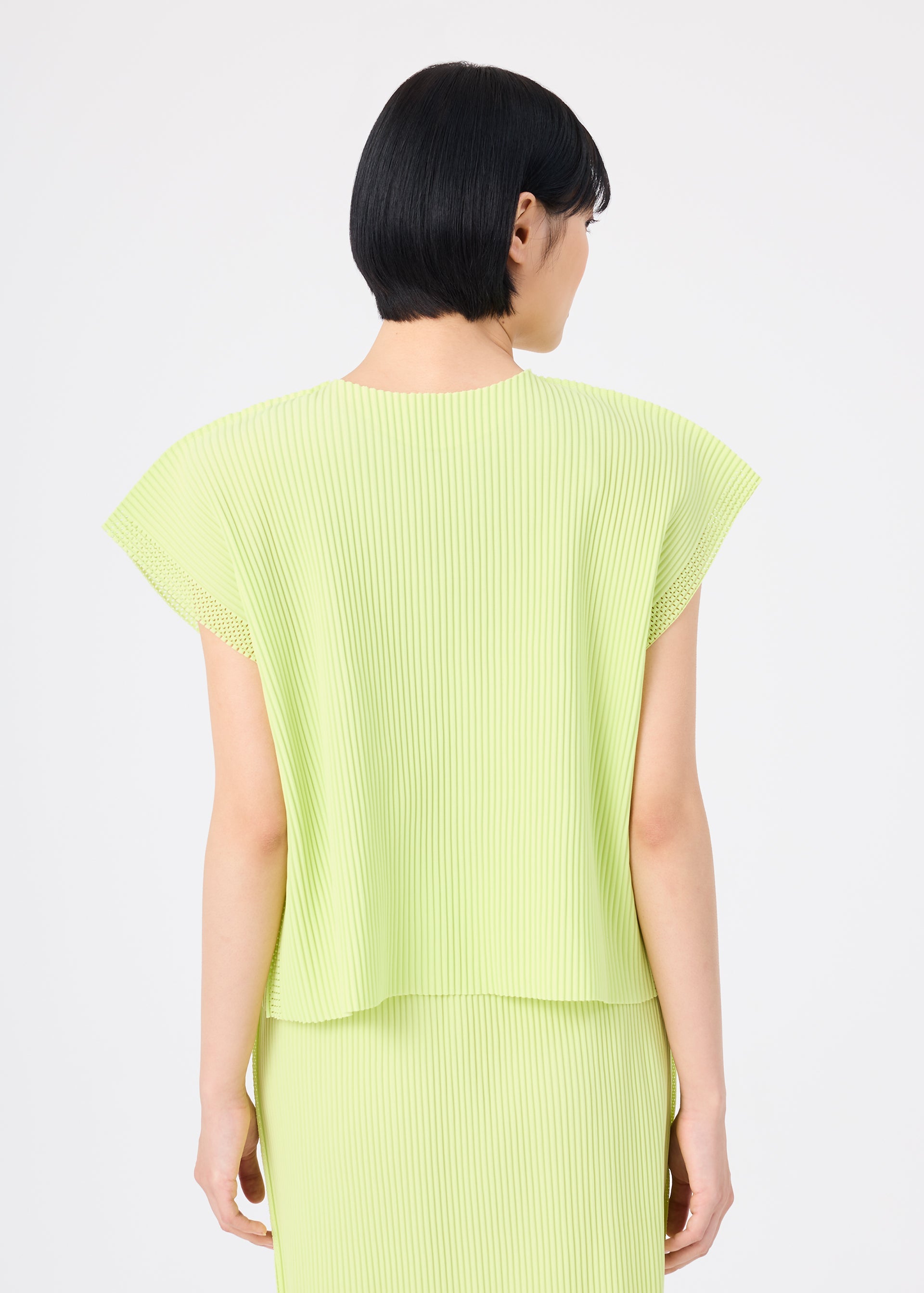 Zoomed view A person with short black hair, seen from behind, wears the light green MESH PLEATS top and matching skirt by me ISSEY MIYAKE, featuring fine knit pleats, against a plain white background.