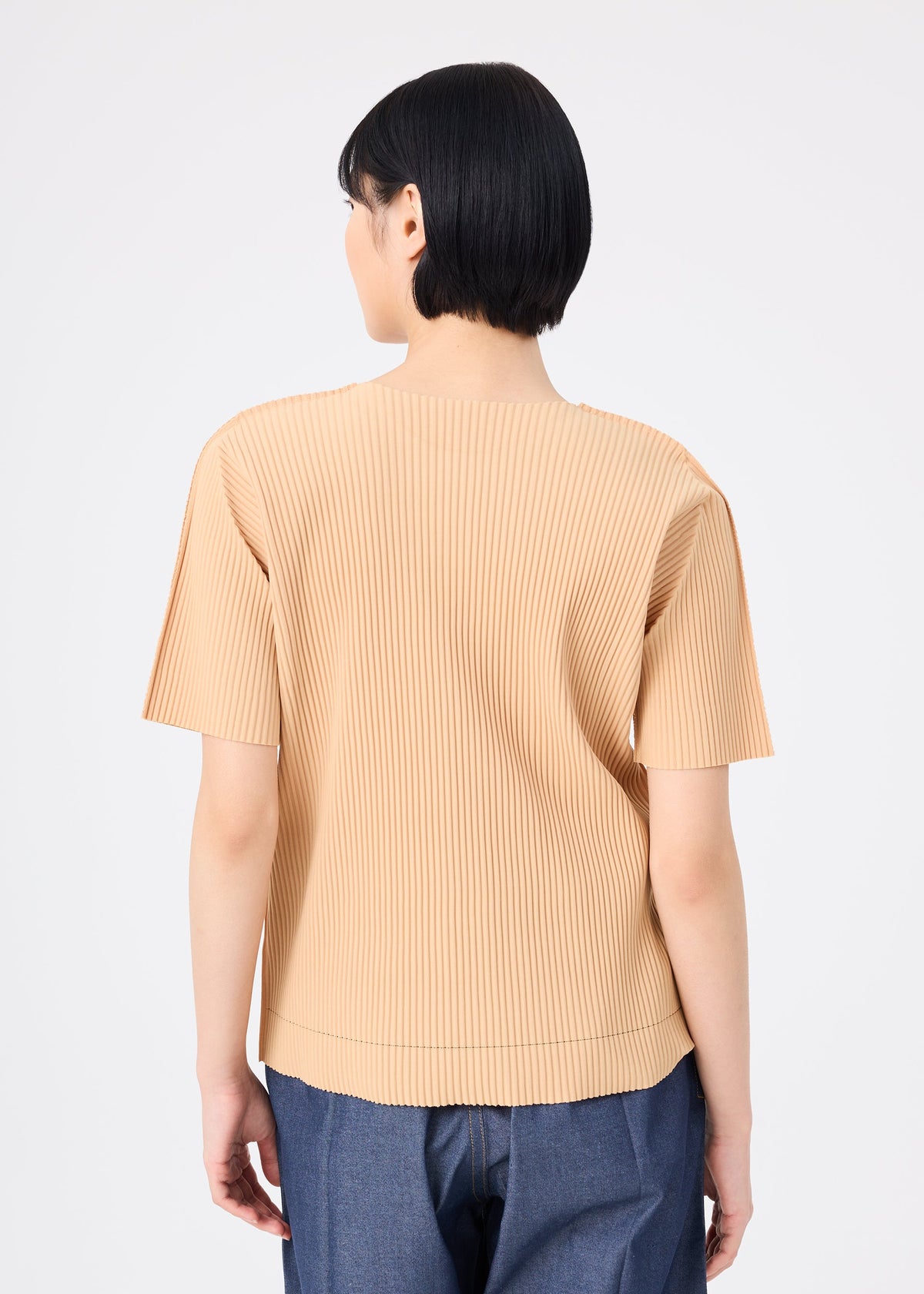 A person with short black hair is seen from the back wearing me ISSEY MIYAKE’s FINE KNIT PLEATS COLOR 1 top—a beige, round-neck, short-sleeved style with fine knit pleats—paired with blue jeans against a plain white background. view 7 of 8