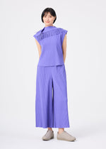 A woman with short black hair stands against a white background wearing the LEAF PLEATS set by me ISSEY MIYAKE—a lightweight lavender ensemble featuring a ruffled top and matching wide-leg pants. She completes the look with beige slip-on shoes.