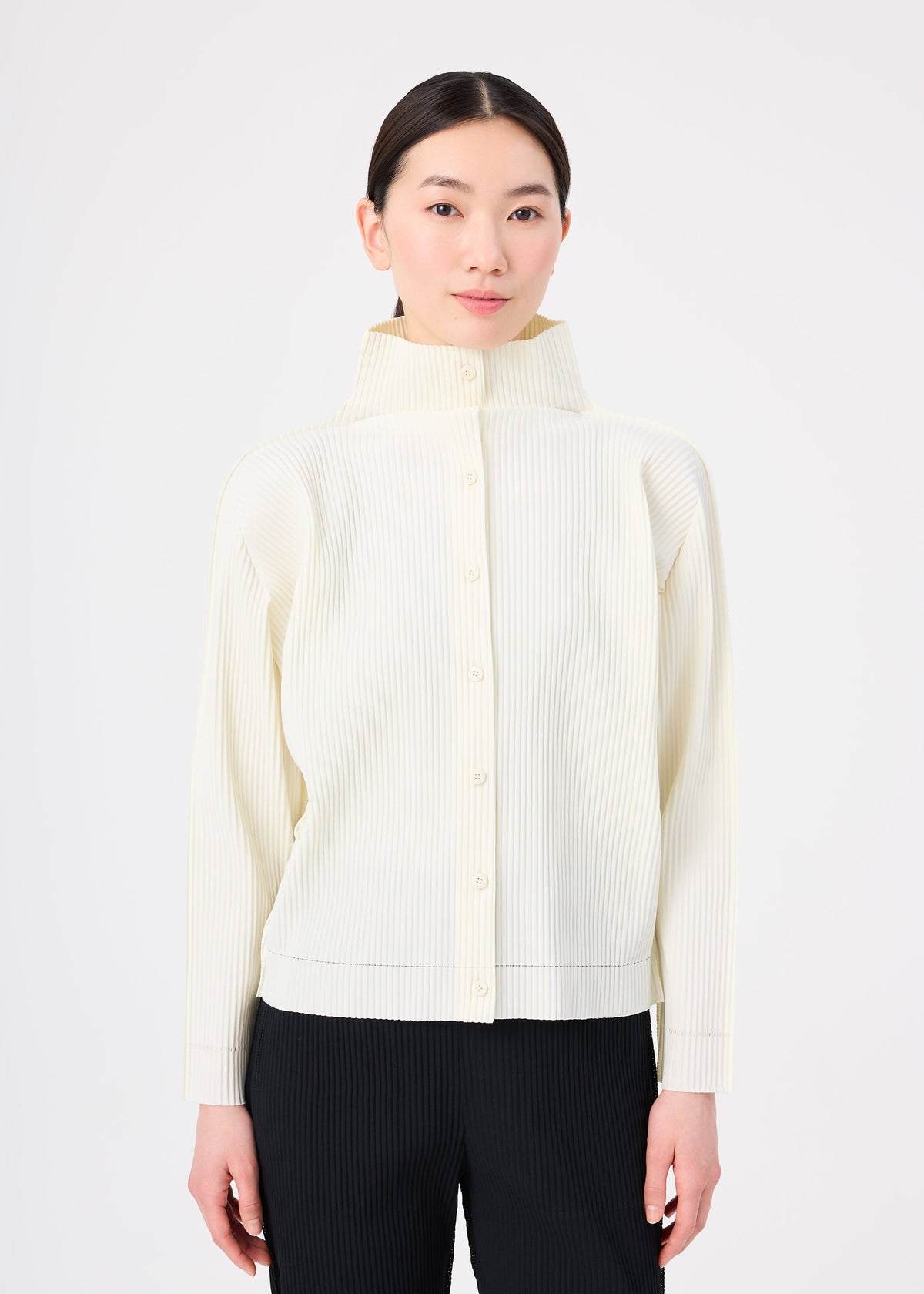 A woman with straight dark hair tied back models the me ISSEY MIYAKE FINE KNIT PLEATS cream high-neck cardigan with black pants against a plain white background. view 4 of 6