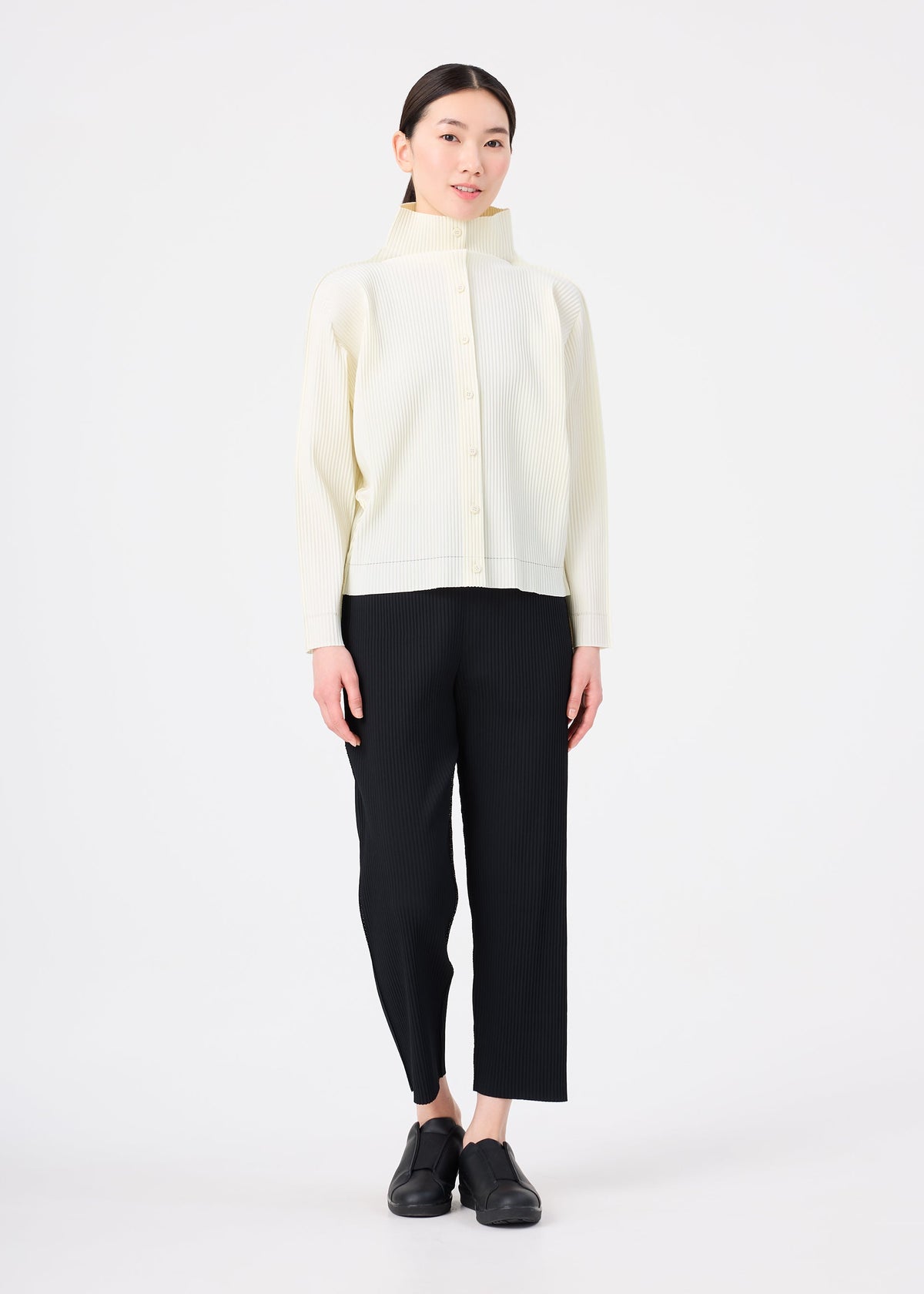 A woman stands against a white background wearing the FINE KNIT PLEATS cardigan by me ISSEY MIYAKE over black straight-leg pants and black shoes, her dark hair tied back and arms relaxed, creating an understated, elegant look. view 6 of 6