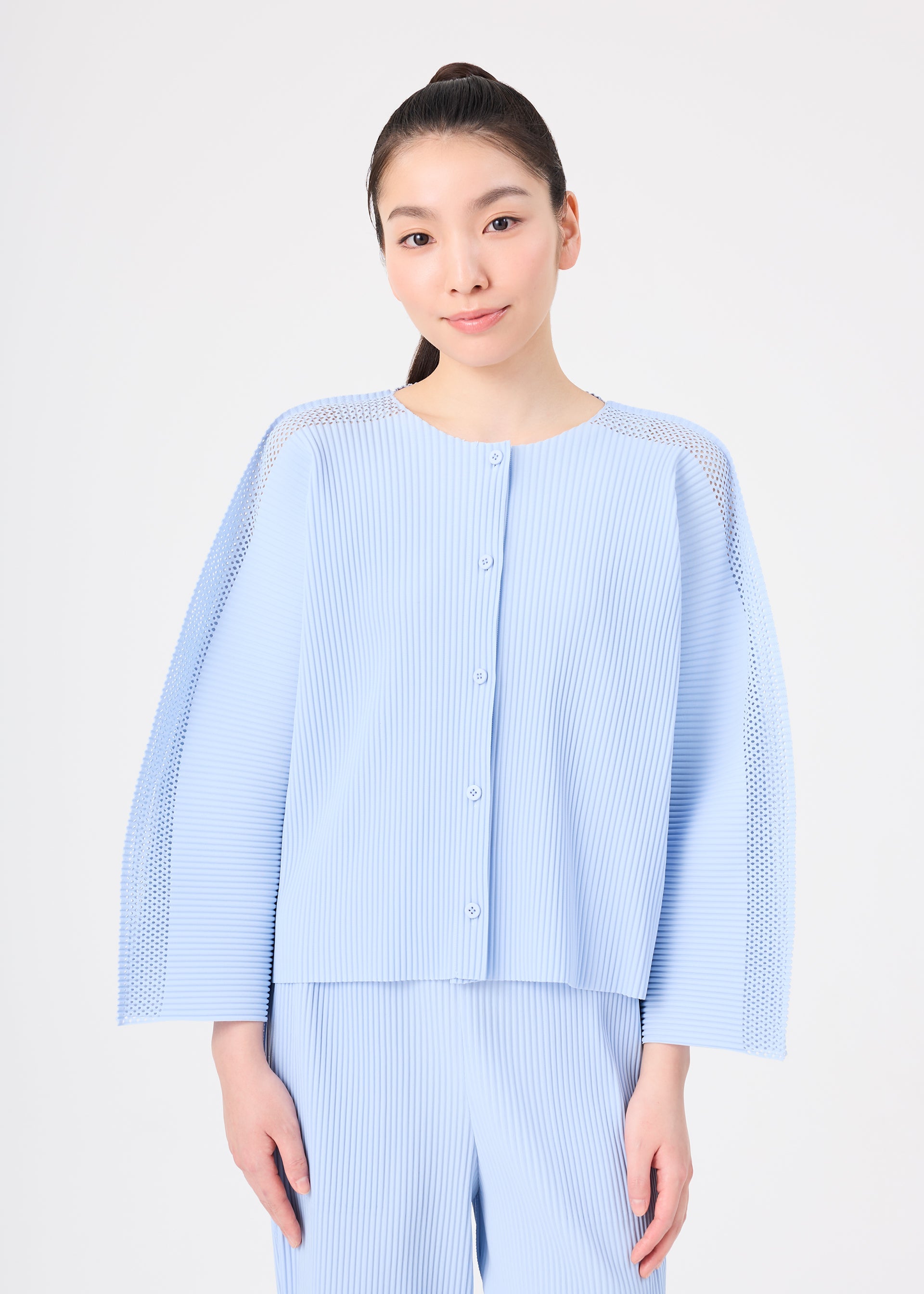 Zoomed view A woman with long dark hair tied back wears the light blue MESH PLEATS set from me ISSEY MIYAKE, featuring a pleated cardigan and matching pants. She stands against a plain white background, smiling gently at the camera.