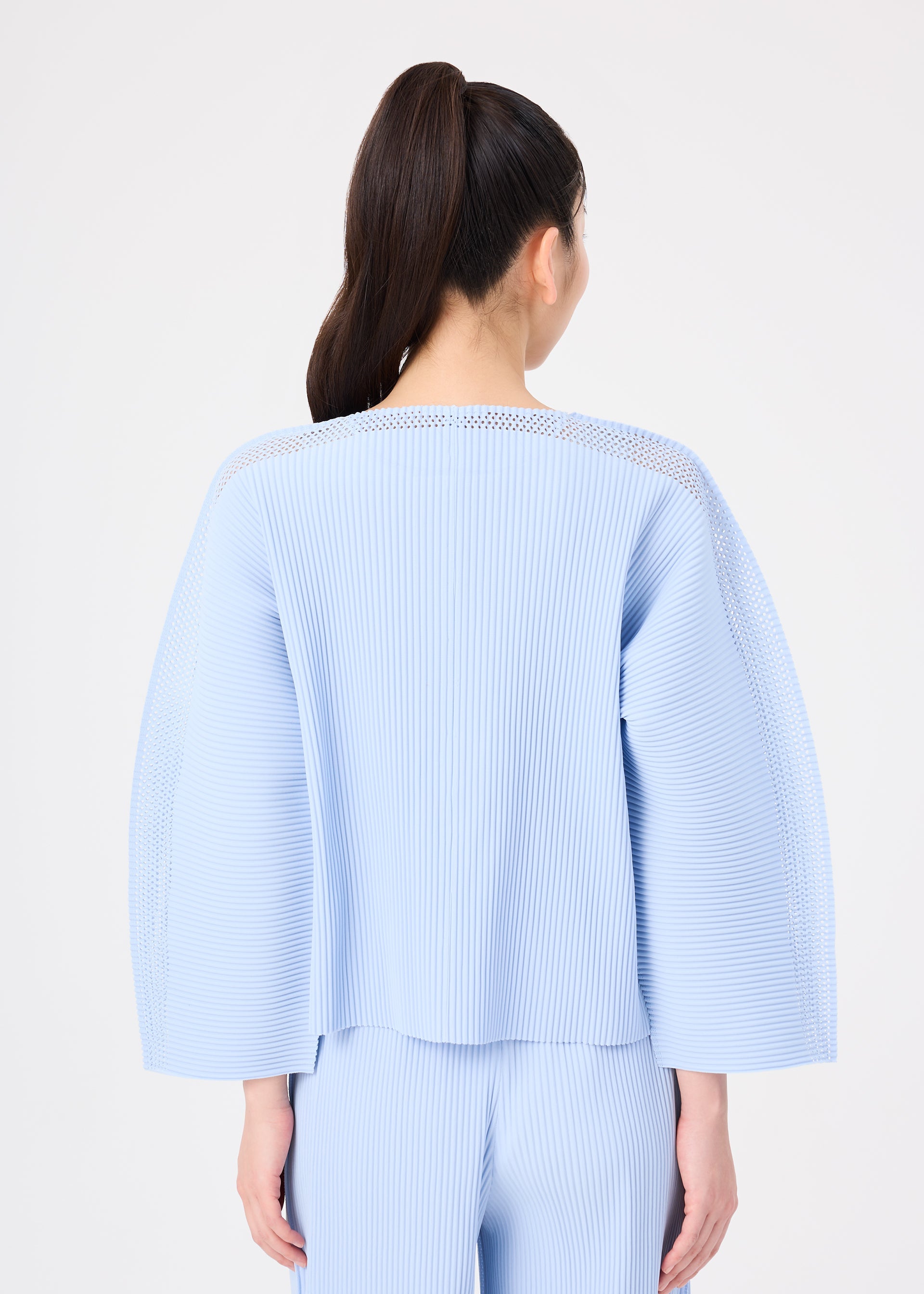 Zoomed view A person with long dark hair in a ponytail is seen from behind, wearing the MESH PLEATS cardigan and matching pants by me ISSEY MIYAKE. The light blue, wide-sleeved pleated set contrasts against a plain white background.