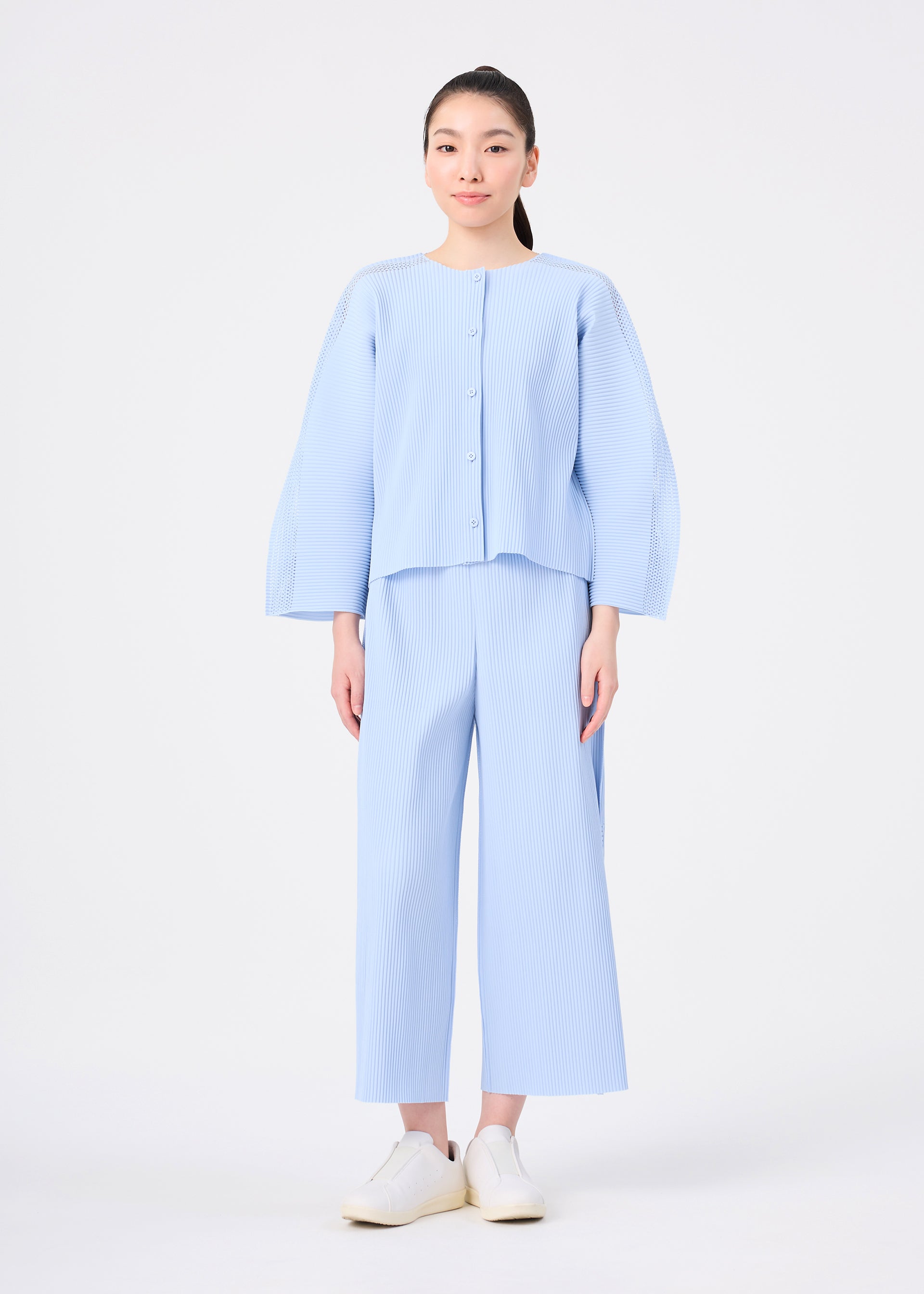 Zoomed view A person stands against a white background wearing the me ISSEY MIYAKE MESH PLEATS light blue cardigan and matching wide-leg pants, paired with white shoes.