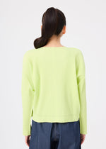A person with long, dark hair in a high ponytail is seen from behind wearing the me ISSEY MIYAKE FINE KNIT PLEATS COLOR 1 cardigan in light yellow and loose dark blue pants against a plain white background.