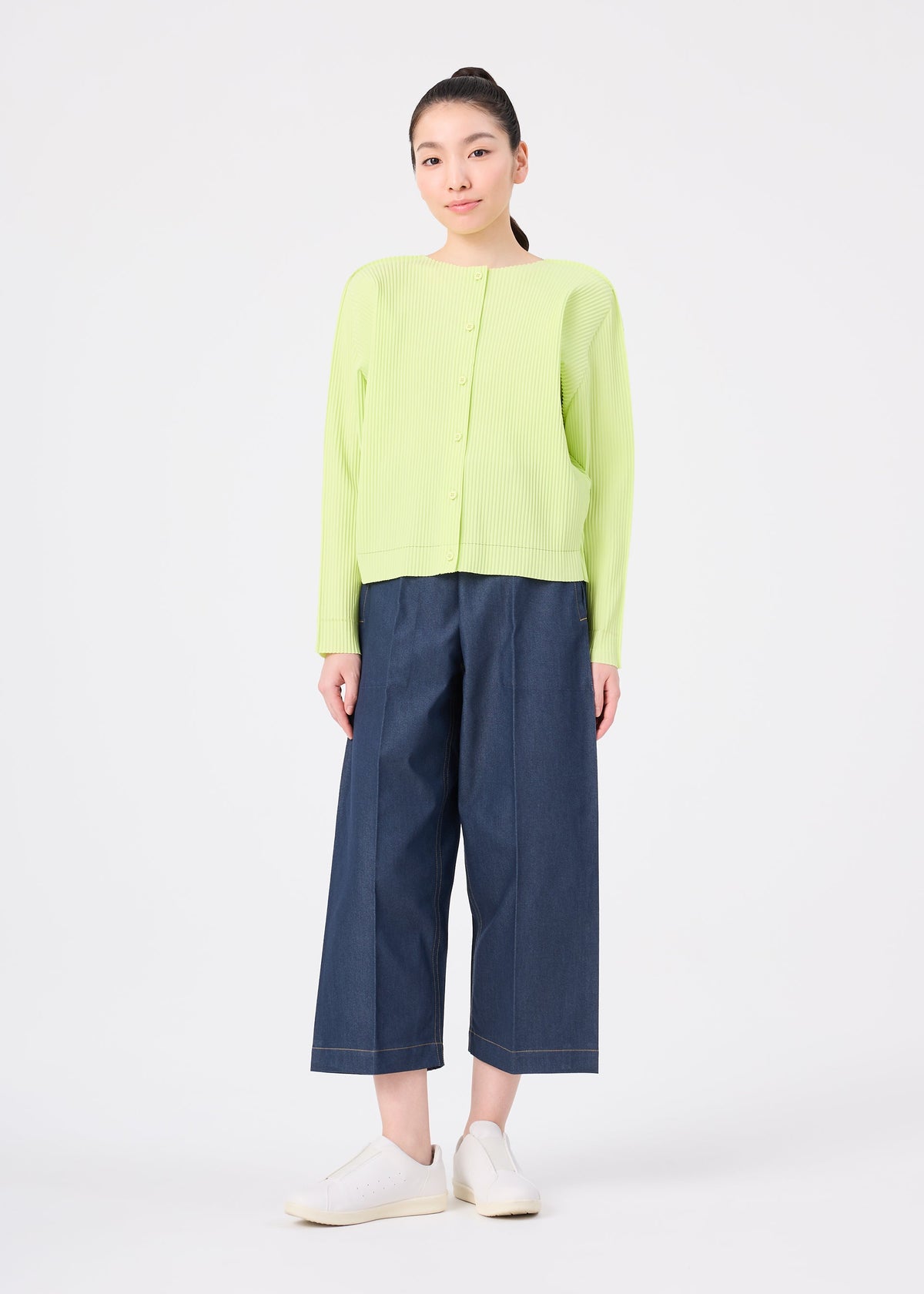 A woman faces forward against a white background, wearing the me ISSEY MIYAKE FINE KNIT PLEATS COLOR 1 cardigan in light green, paired with wide-leg dark blue jeans and white slip-on shoes. view 8 of 8