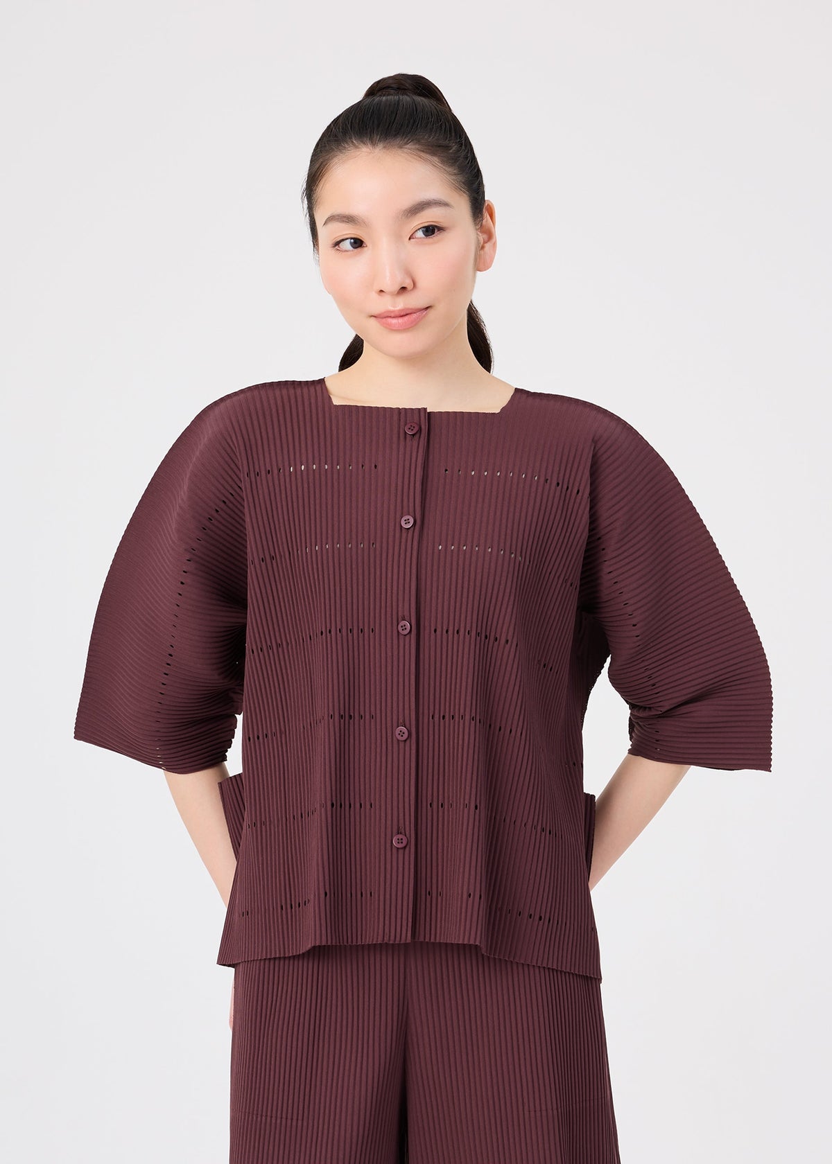 A woman stands against a plain light background, her hair in a ponytail as she gazes slightly to one side. She wears the TOWER PLEATS ensemble by me ISSEY MIYAKE—a burgundy set featuring a pleated top with short, wide sleeves and matching pants. The subtle texture and distinctive design of the outfit echo the refined sophistication and effortless elegance characteristic of pleats cardigans. view 5 of 8