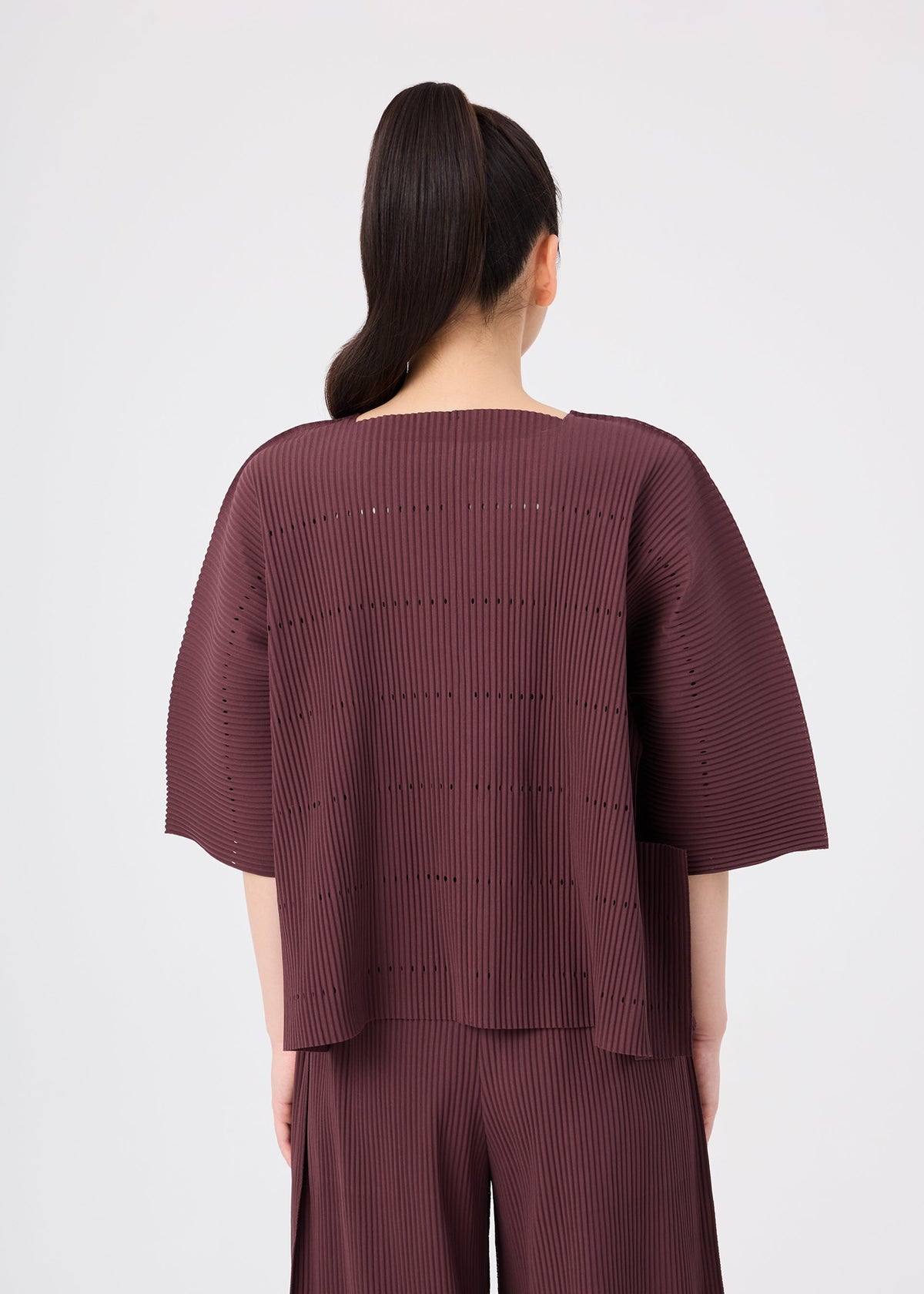 A person with long dark hair in a ponytail is shown from the back against a plain light background, wearing the me ISSEY MIYAKE TOWER PLEATS set—a loose, burgundy pleated top and matching pants—layered with a fine knit cardigan. view 6 of 8