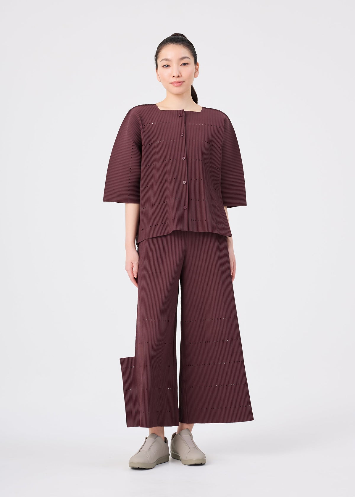A woman stands facing forward, dressed in the me ISSEY MIYAKE TOWER PLEATS ensemble—a matching loose-fitting maroon top and wide-leg pants with subtle stripes—layered beneath a fine knit cardigan for an added touch of softness. She has her hair tied back and is wearing beige shoes, set against a plain white background. view 7 of 8