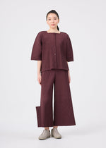 A woman stands facing forward, dressed in the me ISSEY MIYAKE TOWER PLEATS ensemble—a matching loose-fitting maroon top and wide-leg pants with subtle stripes—layered beneath a fine knit cardigan for an added touch of softness. She has her hair tied back and is wearing beige shoes, set against a plain white background.