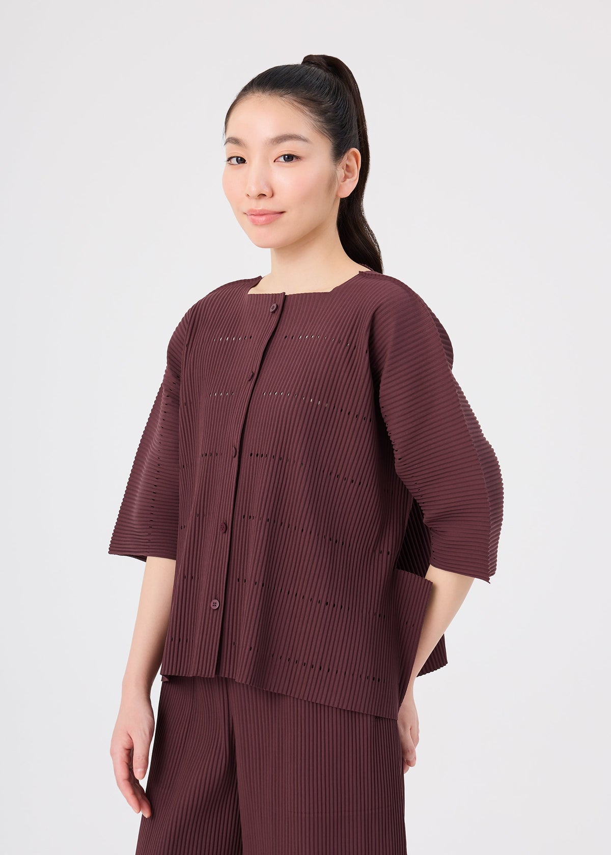 A woman with long dark hair styled in a ponytail, wearing the TOWER PLEATS set by me ISSEY MIYAKE—a loose, pleated, dark burgundy top layered with a fine knit cardigan and matching pants—stands against a plain light background, gazing slightly to the side with a gentle smile. view 8 of 8