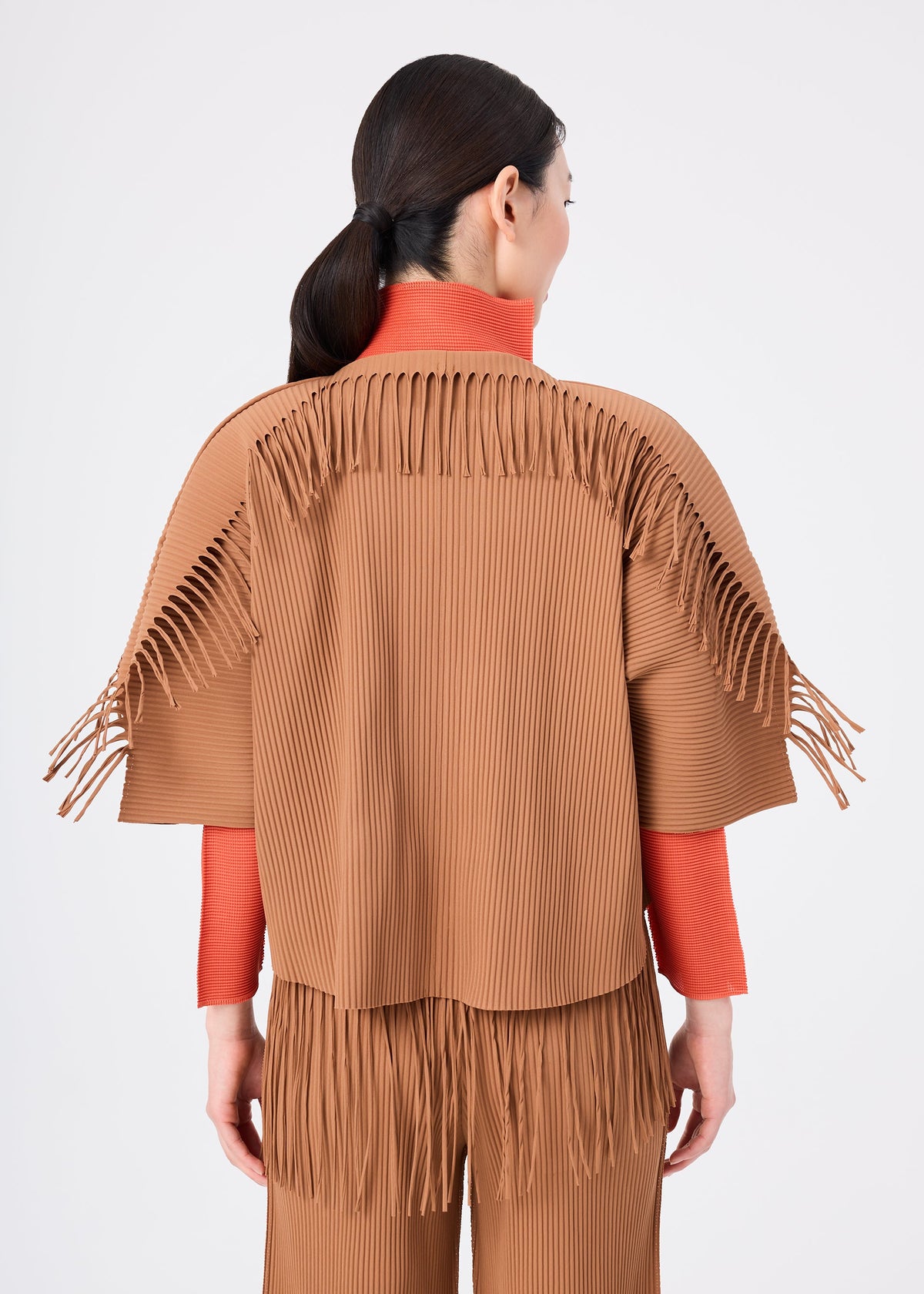 A woman stands facing away, wearing the LEAF PLEATS brown pleated cardigan jacket with fringe on the shoulders and back by me ISSEY MIYAKE, layered over orange sleeves, paired with matching LEAF PLEATS pleated pants featuring fringe at the hips. view 6 of 7