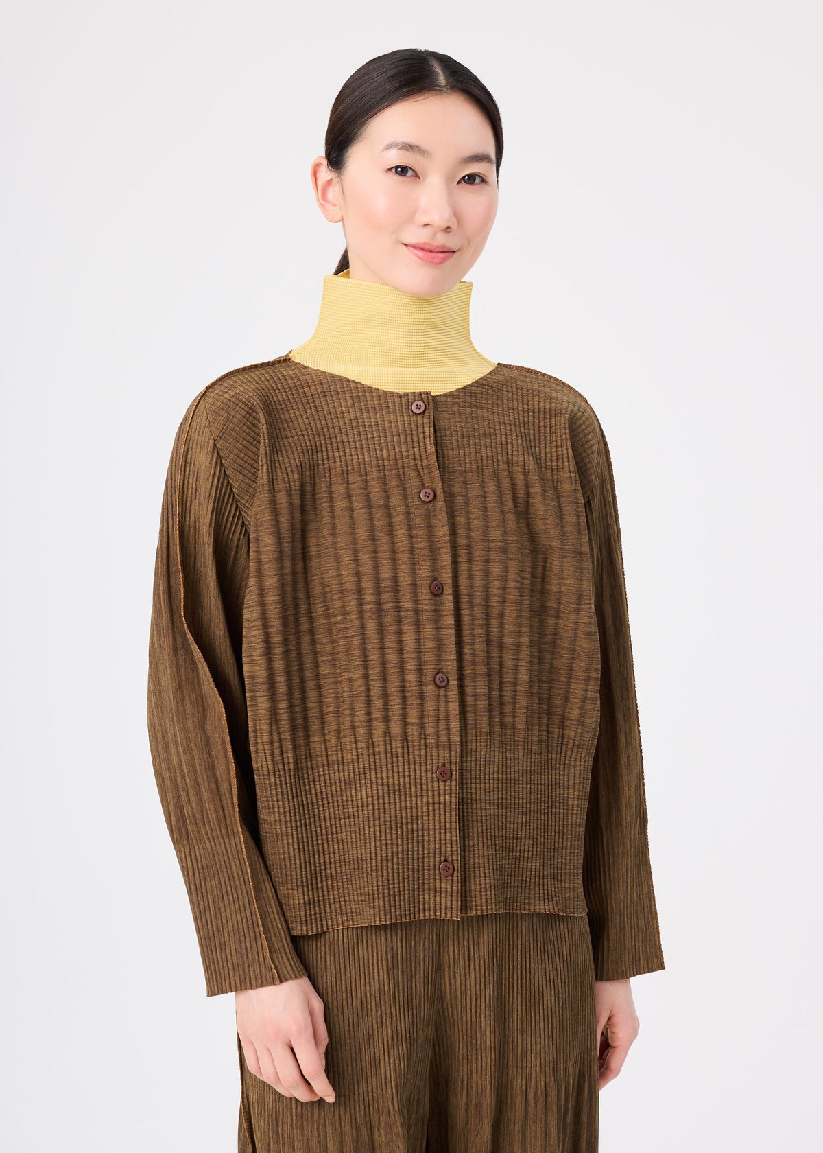 Zoomed view Wearing the MIX FINE KNIT PLEATS by me ISSEY MIYAKE, a person with dark hair tied back pairs the textured brown cardigan and pants with a high-neck yellow shirt against a plain white background.