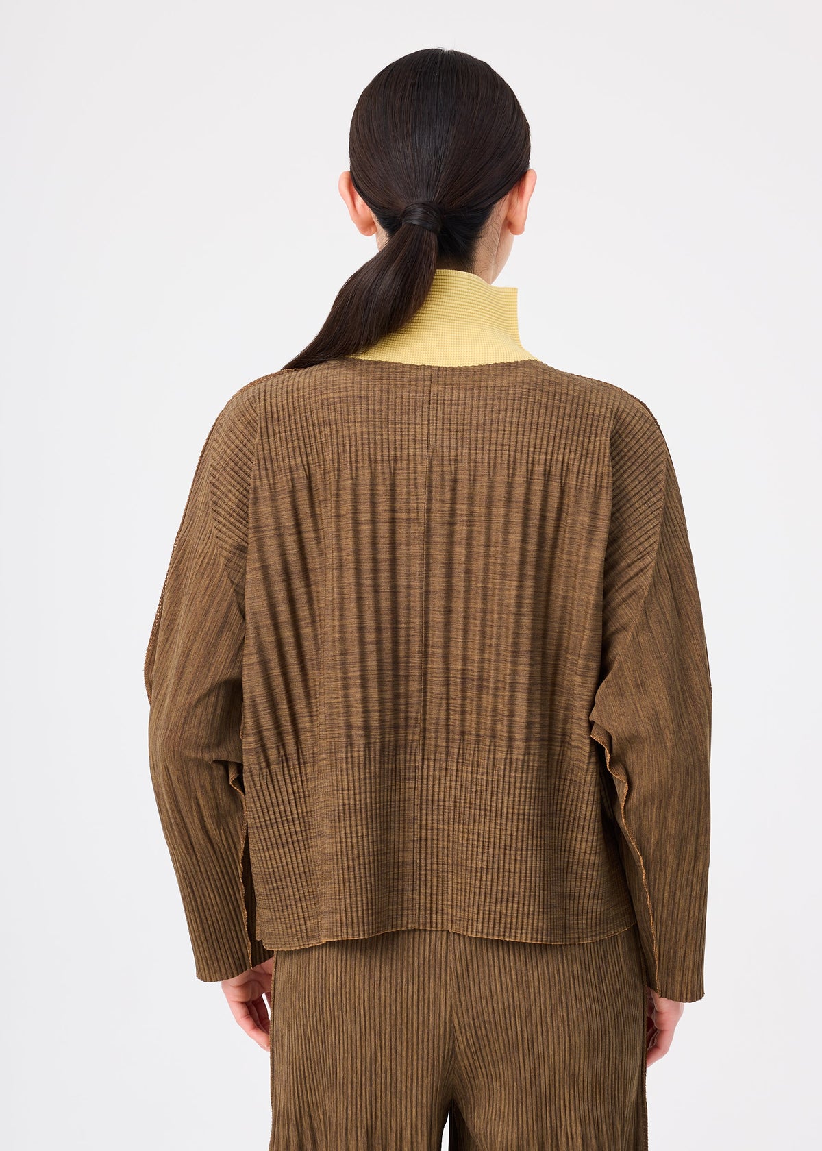 A person faces away, wearing the me ISSEY MIYAKE MIX FINE KNIT PLEATS in brown with matching pants and a yellow turtleneck. Their dark hair is tied back in a low ponytail. view 7 of 8