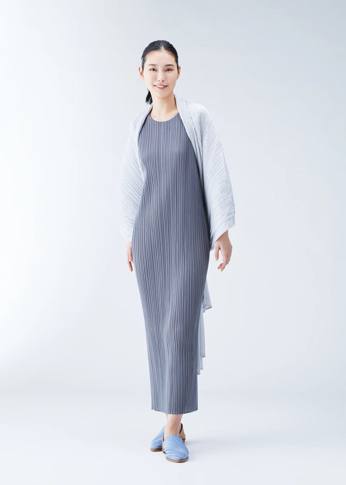 A woman in the BASIC MADAME-T dress by PLEATS PLEASE ISSEY MIYAKE stands against a white background, smiling with a versatile light stole over her shoulders and blue slip-on shoes. view 5 of 6