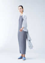 A woman poses against a plain background wearing the PLEATS PLEASE ISSEY MIYAKE BASIC MADAME-T, a loose, sleeveless striped jumpsuit paired with a flowing pleated stole and blue flats, gazing calmly into the camera.
