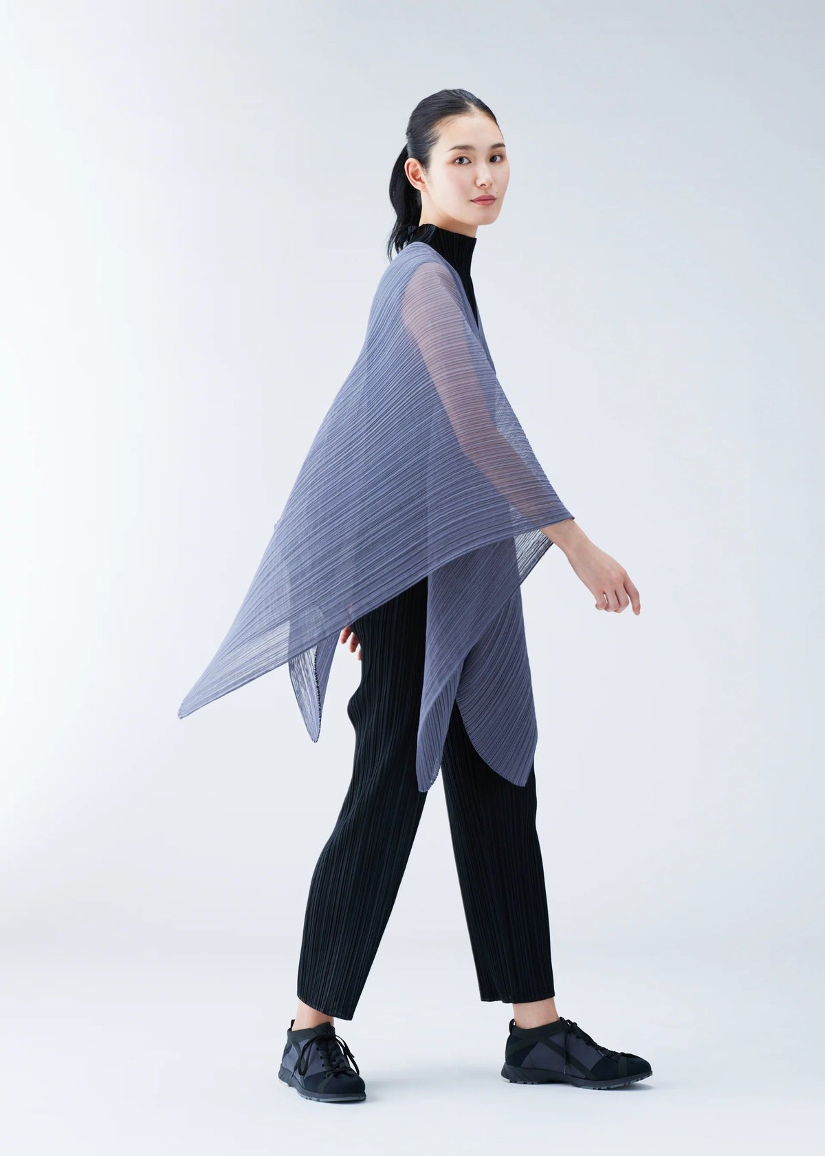 Zoomed view A person wearing black clothes and shoes, styled with the PLEATS PLEASE ISSEY MIYAKE BASIC MADAME-T SMALL, a sheer pleated asymmetrical lavender poncho, stands against a plain light background.