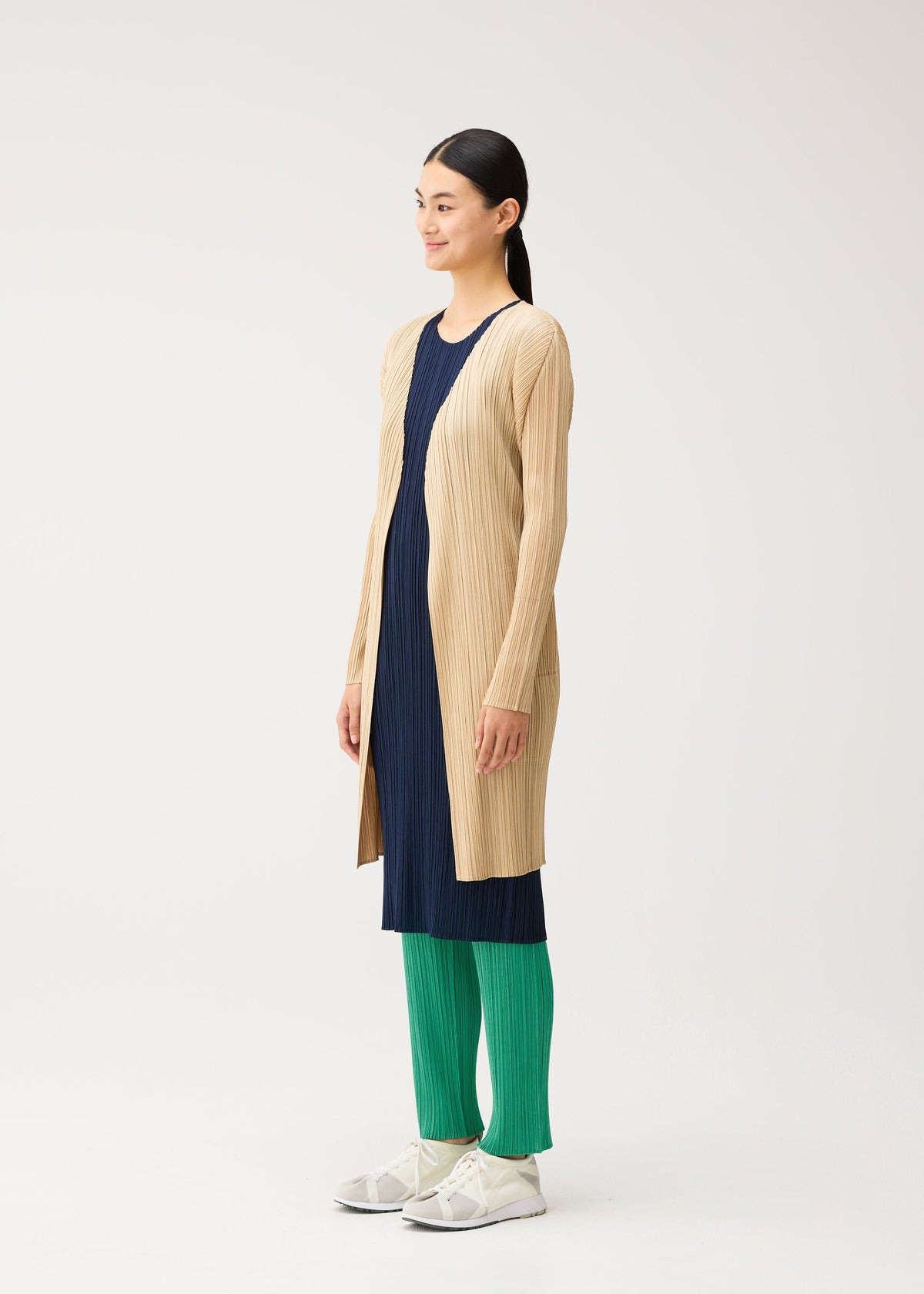Zoomed view A person wears a beige cardigan from the PLEATS PLEASE ISSEY MIYAKE NEW COLORFUL BASICS 5 collection over a dark blue midi dress and bright green trousers, paired with white sneakers.