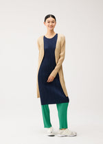 A person wearing a PLEATS PLEASE ISSEY MIYAKE NEW COLORFUL BASICS 5 pleated navy dress, an easy-to-wear beige mid-length coat, and bright green pleated trousers with white sneakers.