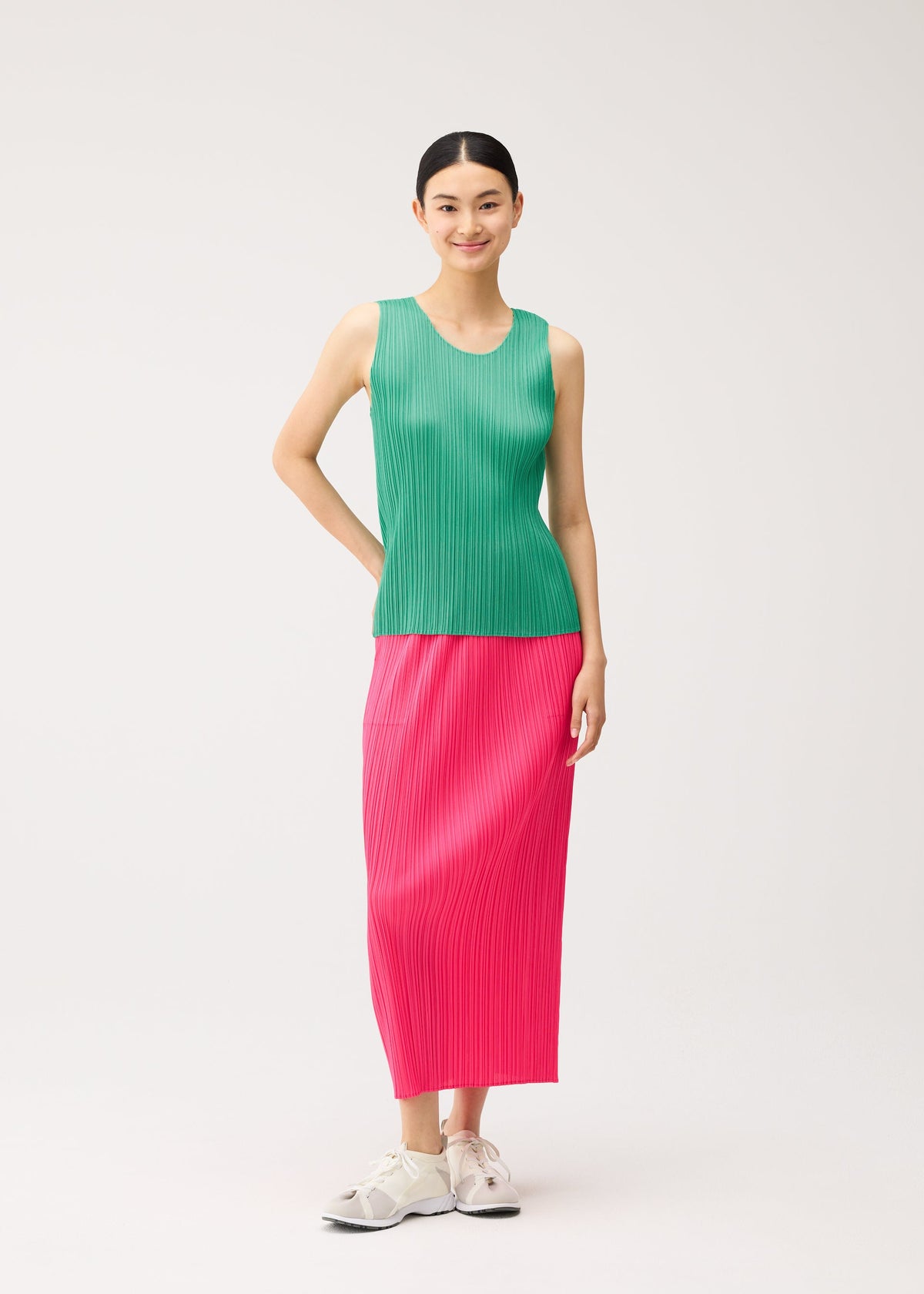 A woman wears a green round-neck sleeveless top from the NEW COLORFUL BASICS 5 collection by PLEATS PLEASE ISSEY MIYAKE, paired with a pink pleated midi skirt and white sneakers. view 10 of 10