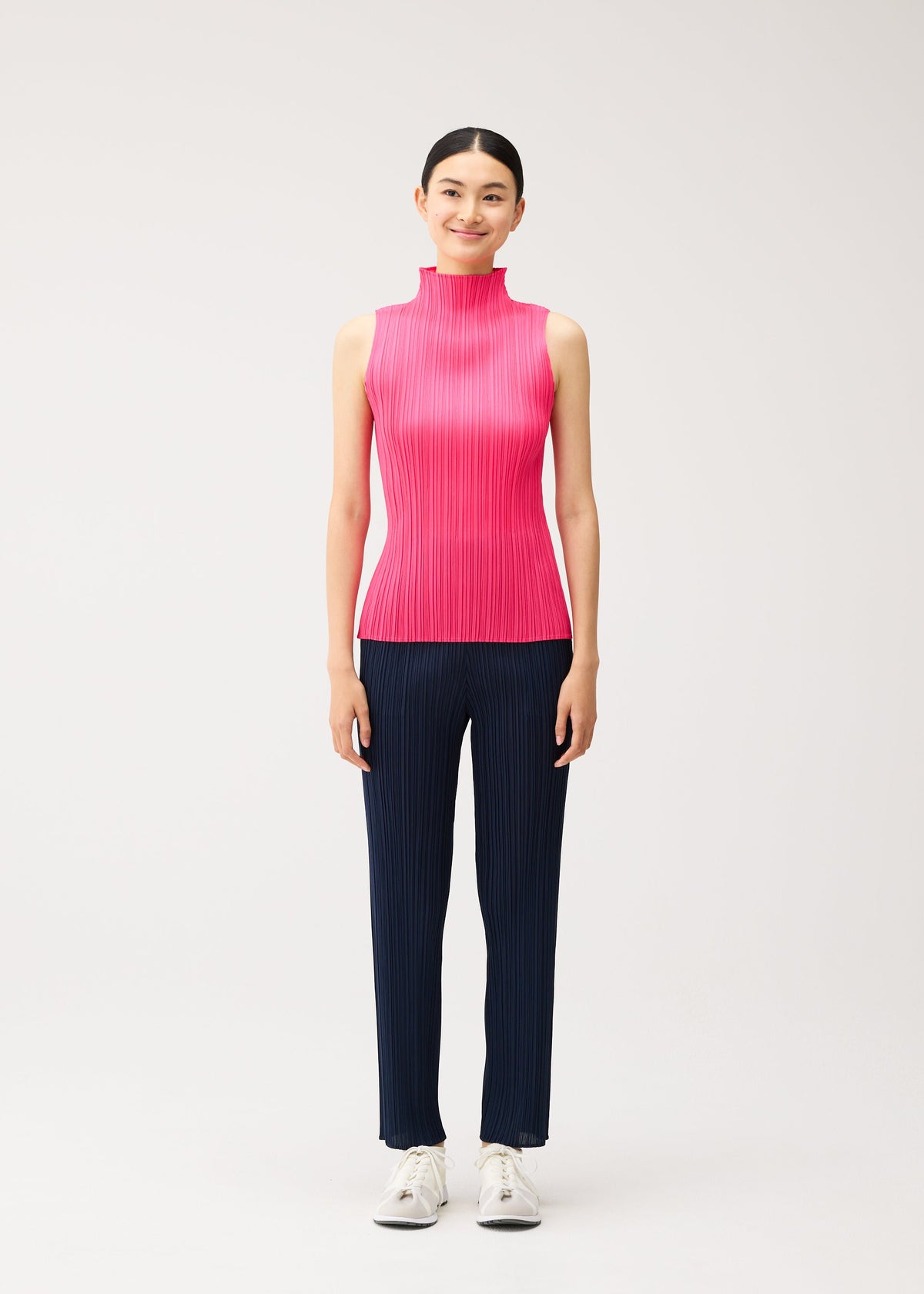 A person wearing a brightly colored, sleeveless mock-neck pleated top and dark blue pleated trousers from the NEW COLORFUL BASICS 5 collection by PLEATS PLEASE ISSEY MIYAKE, finished with white sneakers. view 7 of 10