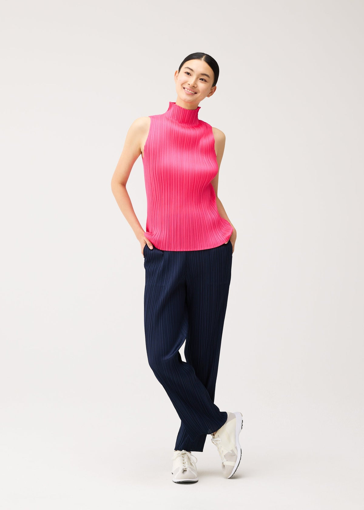 A person wears a NEW COLORFUL BASICS 5 high-neck sleeveless shirt in bright pink with dark blue pleated trousers, standing with their hands in their pockets. view 10 of 10