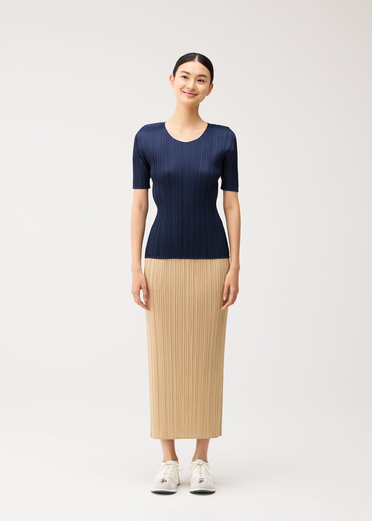 A person wears a navy blue, short-sleeved round-neck NEW COLORFUL BASICS 5 top and a long, tan pleated skirt with white sneakers. view 7 of 10