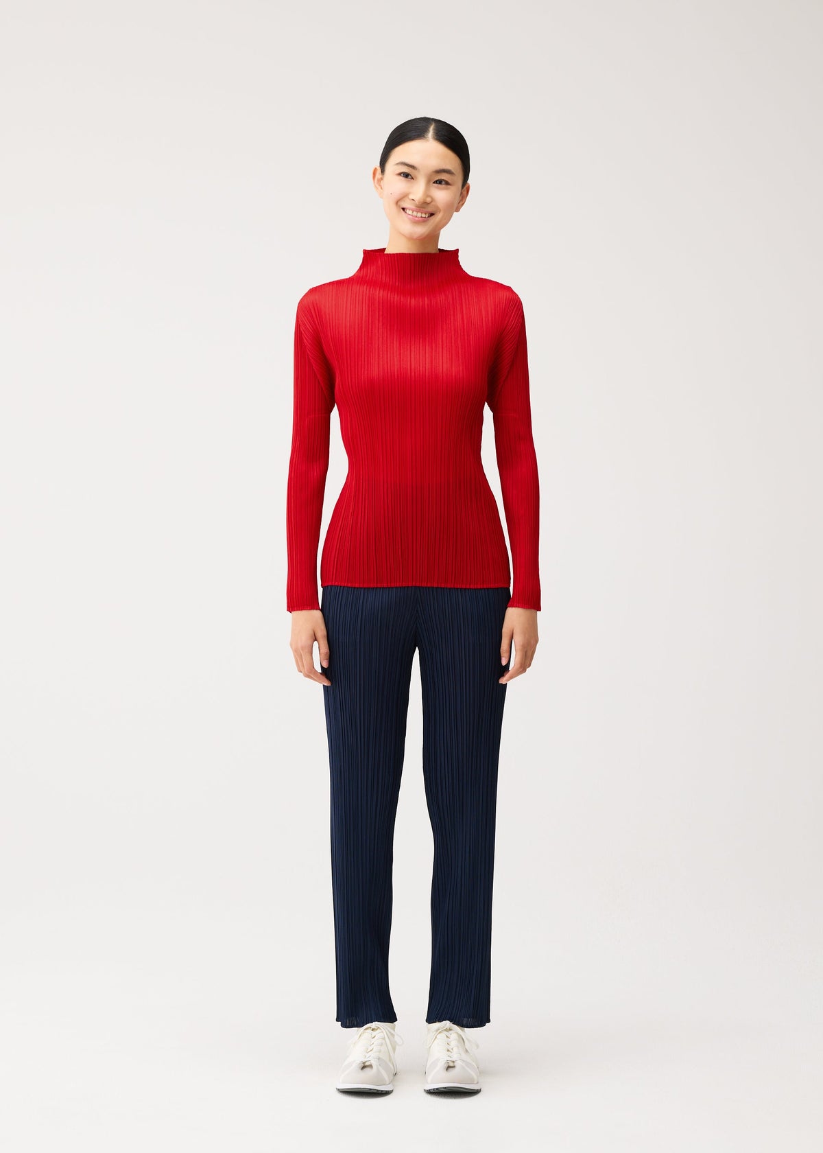 Zoomed view A person wearing a PLEATS PLEASE ISSEY MIYAKE NEW COLORFUL BASICS 5 long-sleeved, brightly colored red pleated mock-neck top and navy pleated trousers with white sneakers.