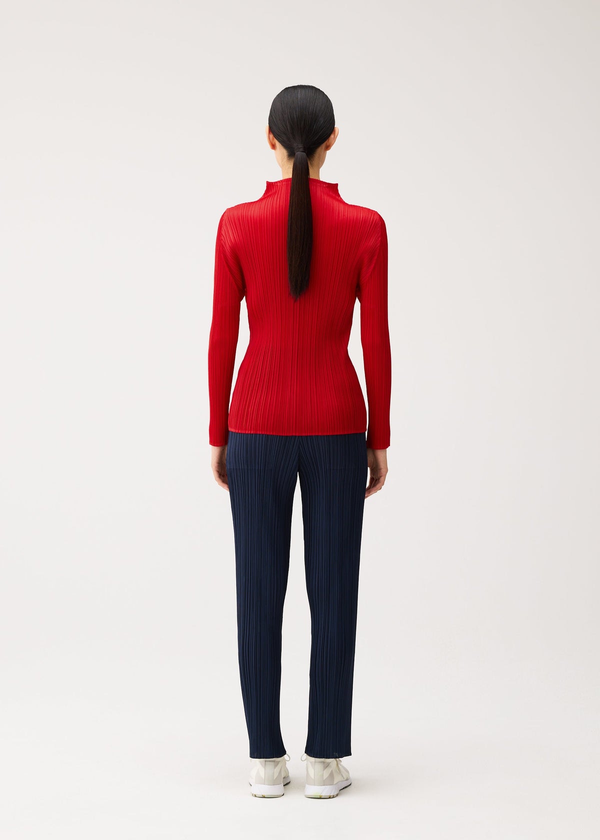 Zoomed view A person viewed from behind wearing a bright, high-neck red long-sleeved pleated top and navy pleated trousers from the NEW COLORFUL BASICS 5 collection by PLEATS PLEASE ISSEY MIYAKE, finished with white sneakers.