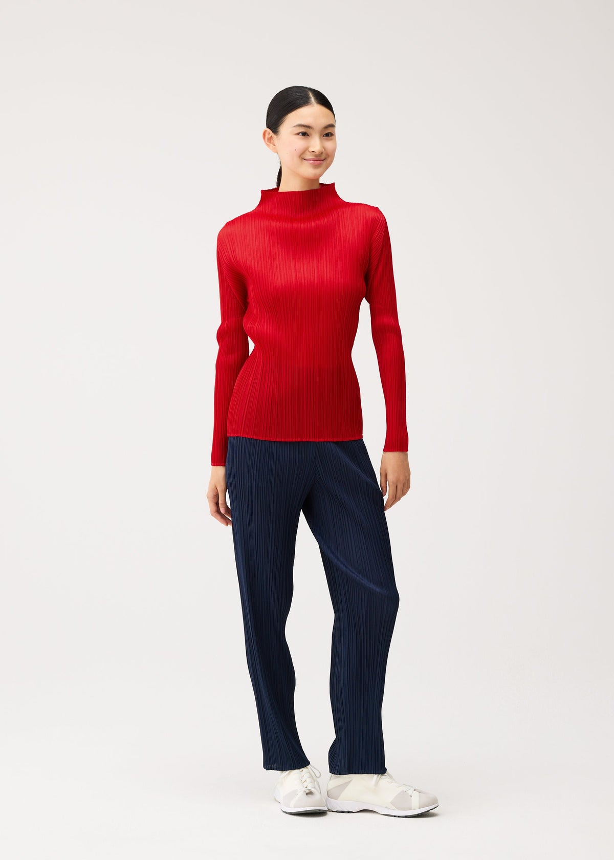 Zoomed view A woman wears a NEW COLORFUL BASICS 5 long-sleeved, high-neck pleated red top and navy pleated trousers, paired with white sneakers.