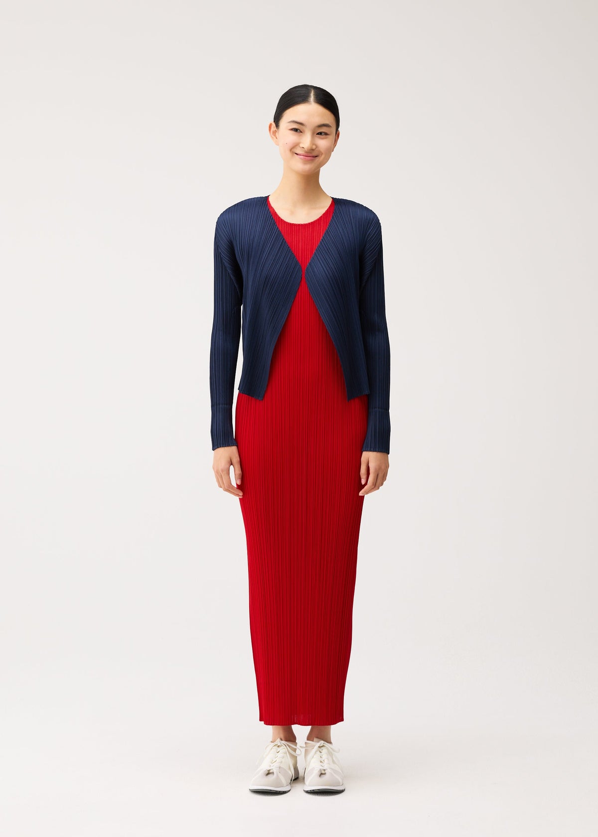 Zoomed view A woman wears a long, brightly colored red sleeveless pleated dress from the NEW COLORFUL BASICS 5 collection by PLEATS PLEASE ISSEY MIYAKE under an easy-to-style navy blue long-sleeved pleated cardigan.
