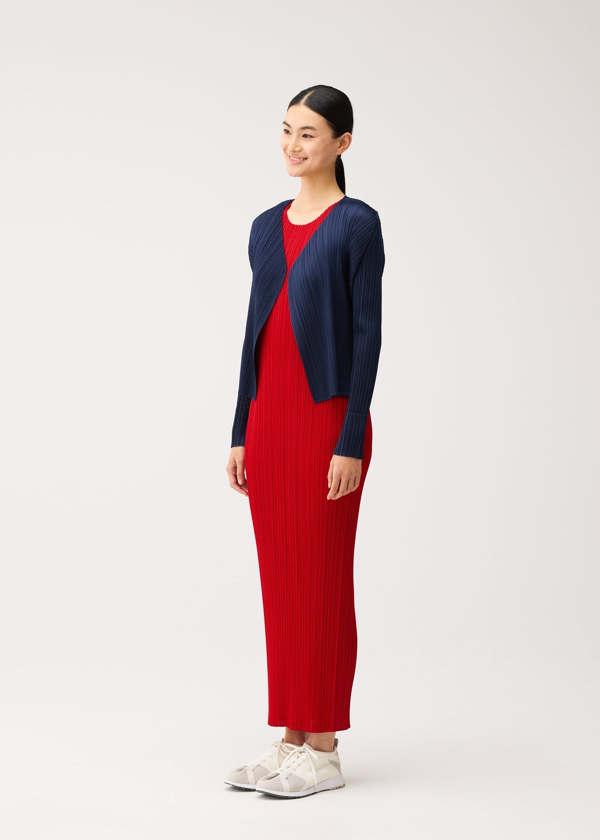 A woman wears a brightly colored red maxi dress from the NEW COLORFUL BASICS 5 collection by PLEATS PLEASE ISSEY MIYAKE, layered under a navy blue pleated short cardigan and paired with white sneakers. view 8 of 10